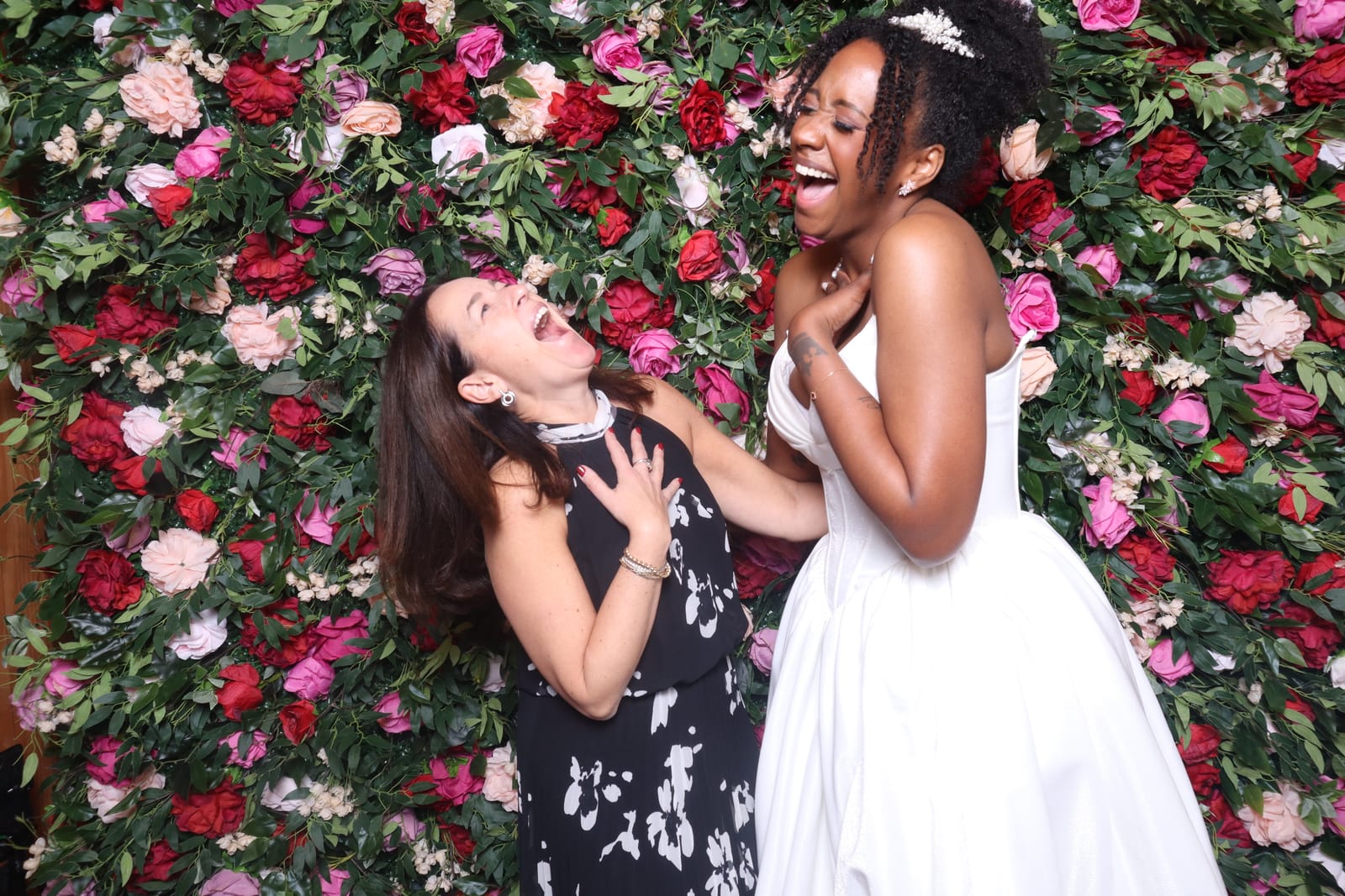 Two friends laughing together, one in a white wedding gown, at the Love Story rose flower wall