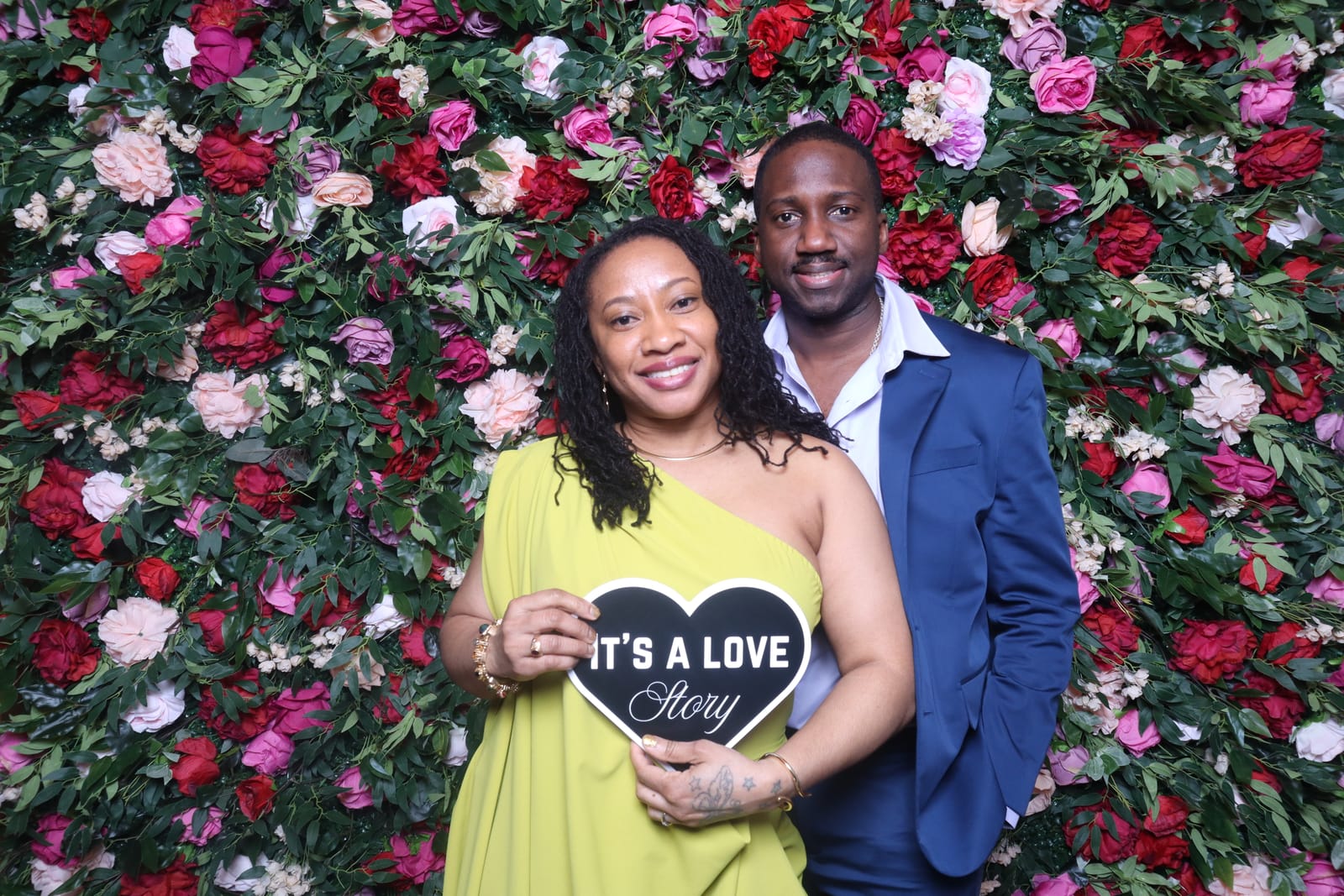 Couple posing with an It's a Love Story heart sign at the rose flower wall