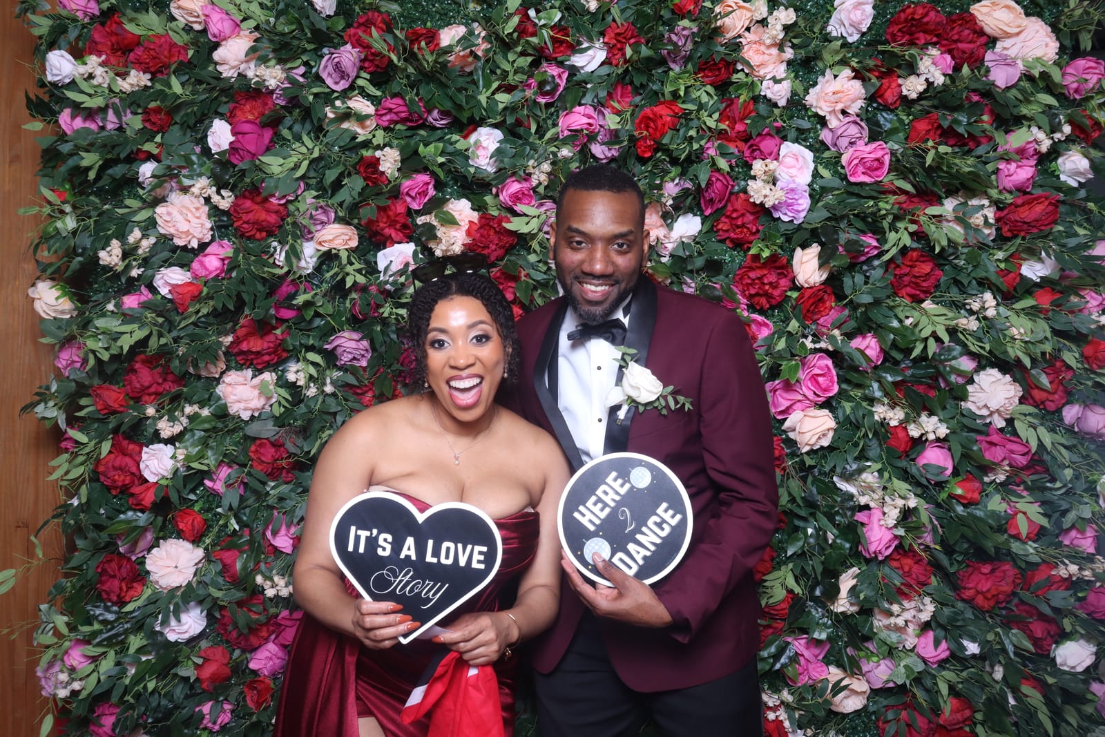 Couple in burgundy posing with It's a Love Story and Here 2 Dance signs at the wedding reception