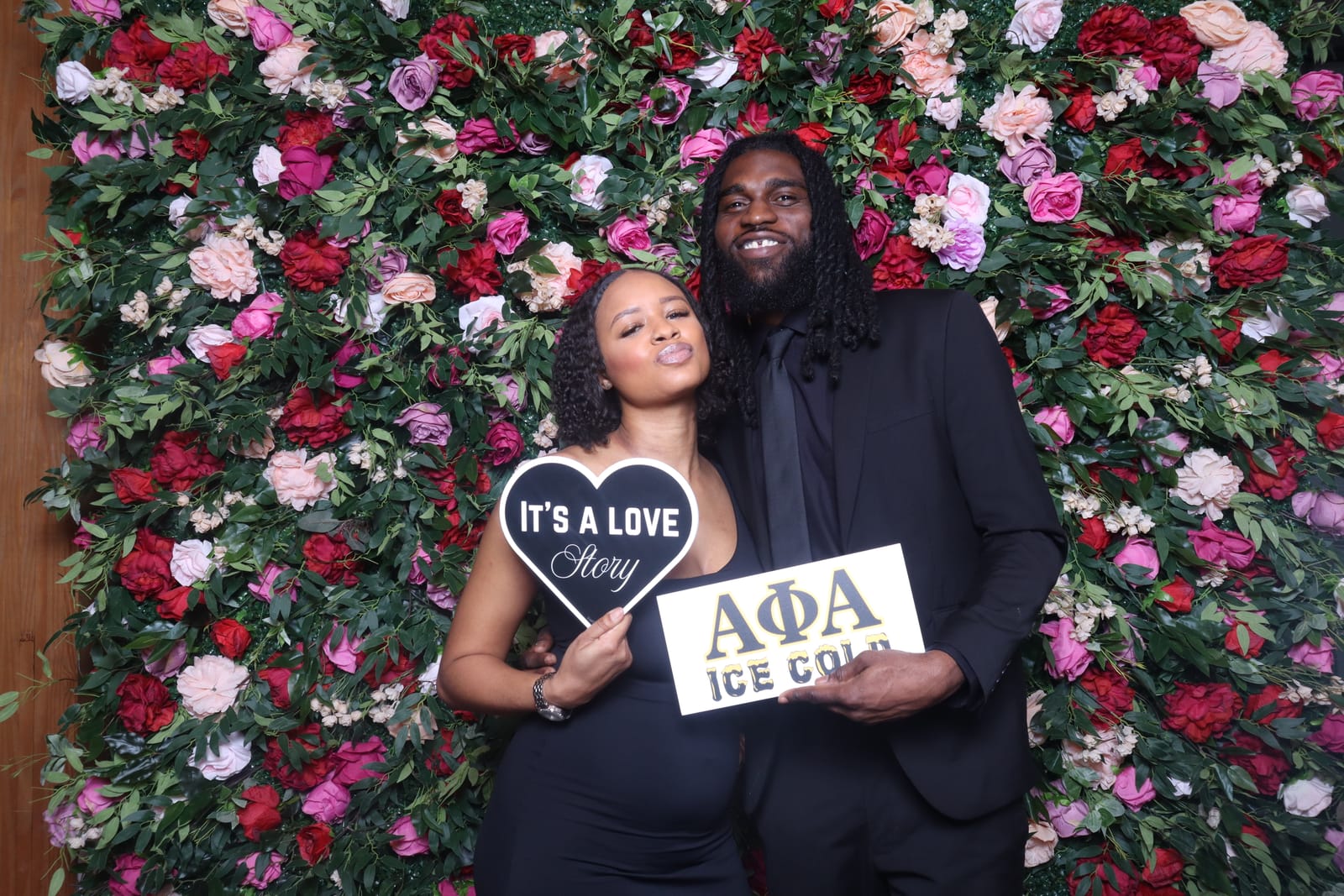 Couple posing with an It's a Love Story heart sign and an Alpha Phi Alpha sign at the reception